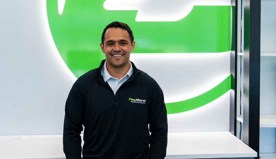 Man wearing black zip-up jacket with PayMore logo standing in front of green and white wall